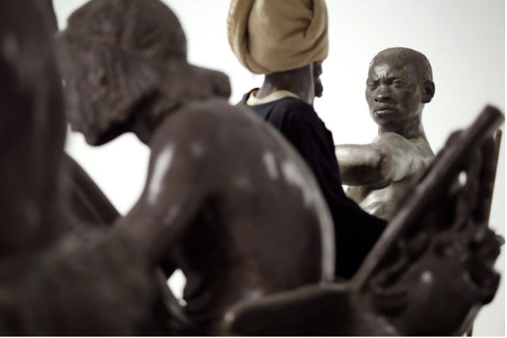 photographie de 4 statues en bronze coloniales montrant des hommes noirs en gros plan. et dont l'un fixe le spectateurice droit dans les yeux depuis le coin supérieur droit de l'image